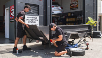 Two mechanics working on a trailer at an approved inspection station with tools and equipment outside a workshop.